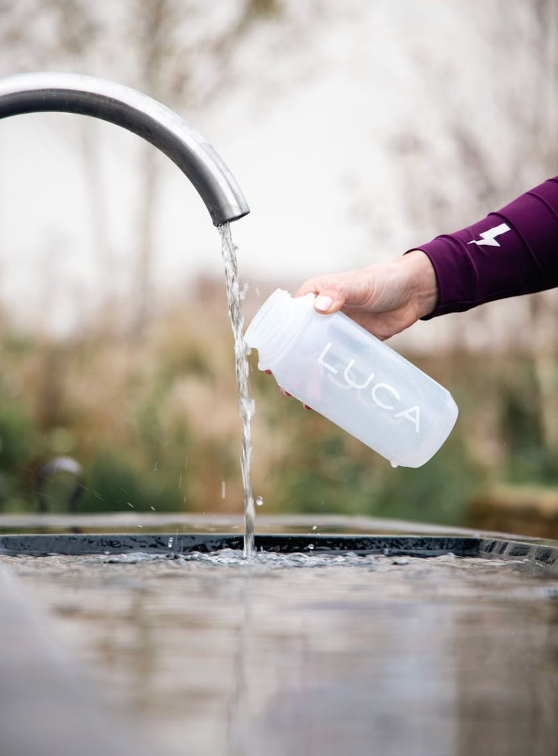 Hand-wearing-a-dark-purple-cycling-jersey-filling-a-translucent- white-LUCA-water-bottle-from-an- outdoor-water-tap