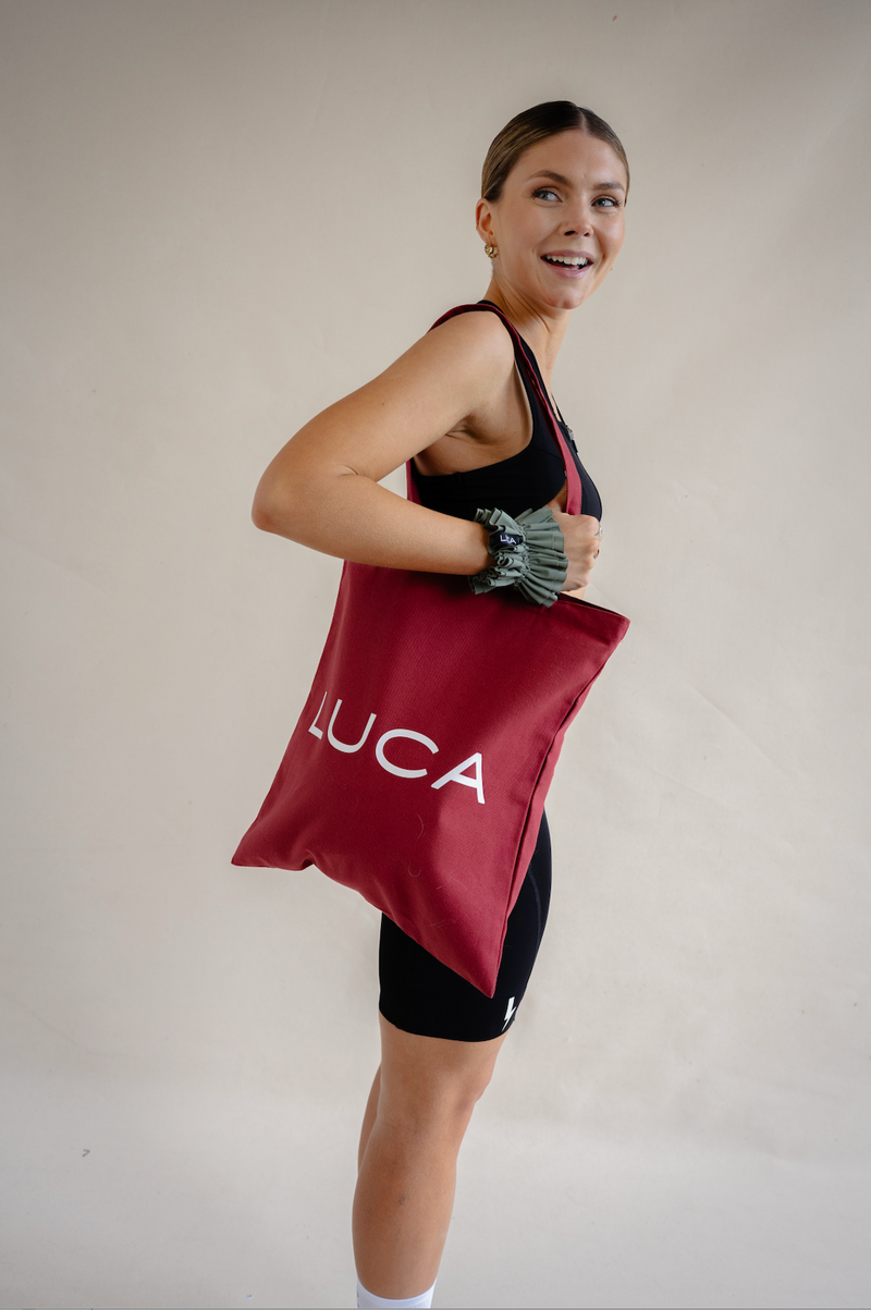 A woman modeling the LUCA® canvas tote bag in burgundy over her shoulder, featuring the white LUCA® logo, paired with black athletic activewear.