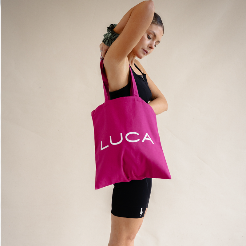A woman modeling a magenta pink LUCA® canvas tote bag over her shoulder, showing the white LUCA® logo in a studio lifestyle shot.