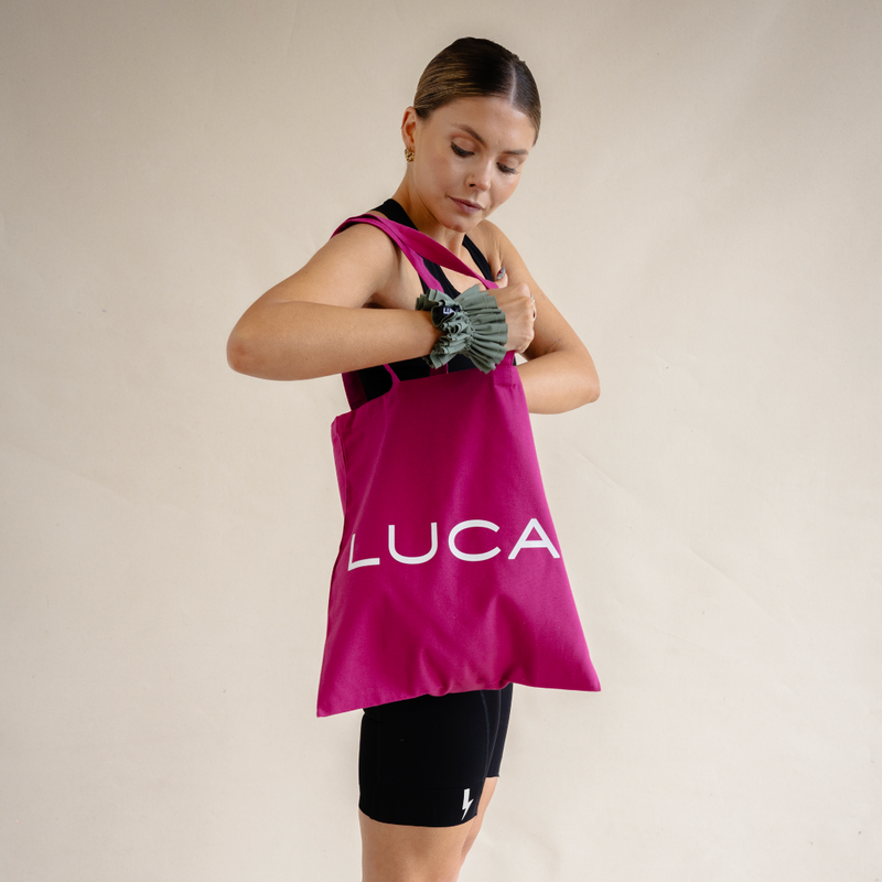A woman modeling a magenta pink LUCA® canvas tote bag over her shoulder, showing the white LUCA® logo in a studio lifestyle shot.
