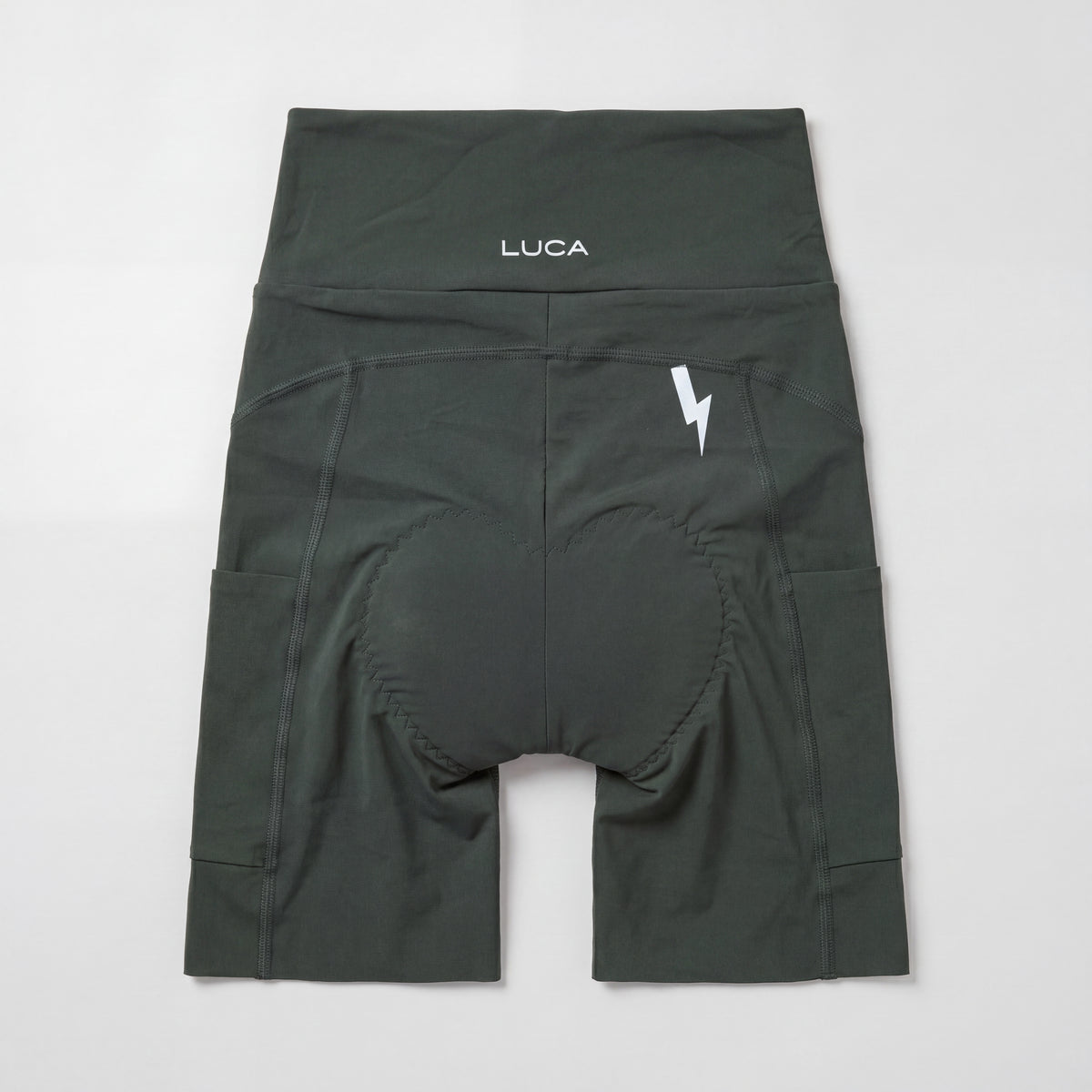 Rear view of a pair of dark basil green cycling bibless shorts centered on a light gray background, showcasing a high waistband with a white LUCA® logo, a white lightning bolt graphic on the upper right side, side pockets, and an integrated padded chamois.