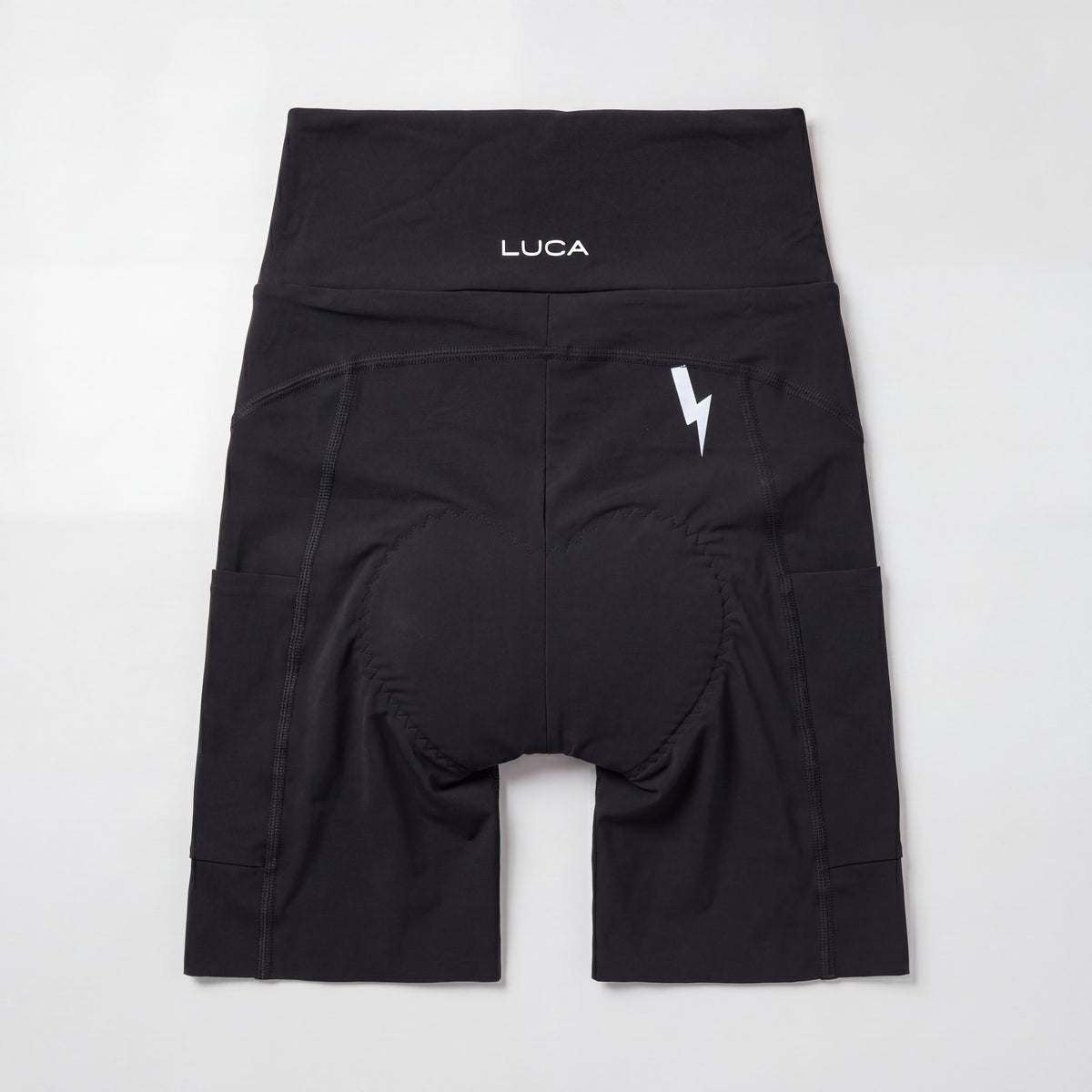 Rear view of a pair of black cycling bibless shorts centered on a light gray background, showcasing a high waistband with a white "LUCA" logo, a white lightning bolt graphic on the upper right side, side pockets, and an integrated padded chamois.