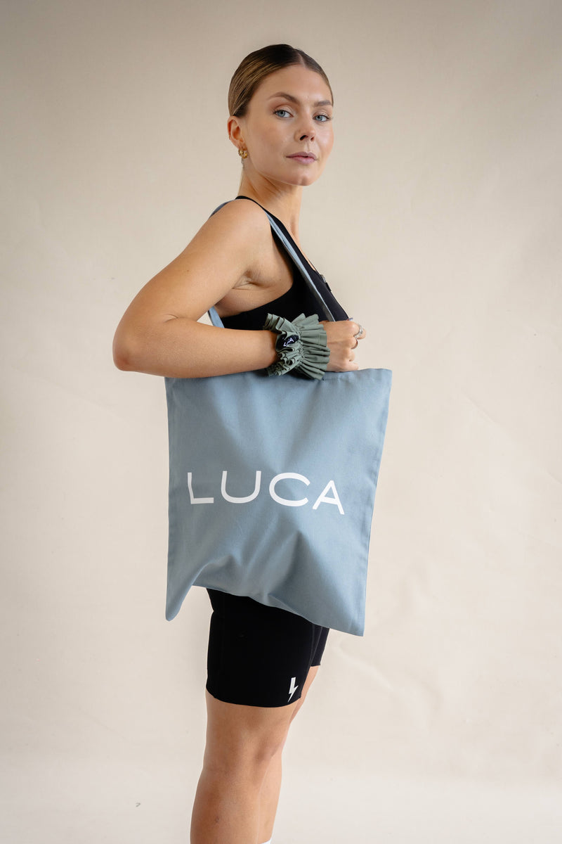 A side profile lifestyle shot of a woman looking over her shoulder against a cream background, holding a slate blue tote bag over her shoulder with a large white LUCA® brand name printed on it. She is wearing a black athletic set, and a matcha green ruffled fabric scrunchie is visible on her wrist.