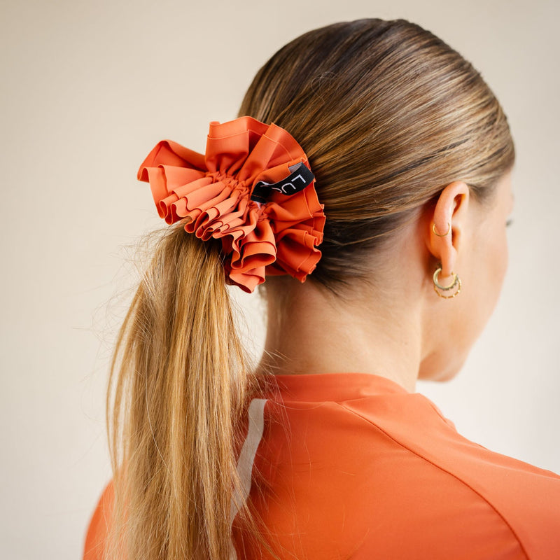 Close-up shot of a person wearing the LUCA Uplift Scrunchie in 'Aperol' orange on their wrist. The accessory features a unique, heavily ruffled tiered fabric design and a small black woven tag displaying the white LUCA logo, paired alongside dark olive green athletic wear.