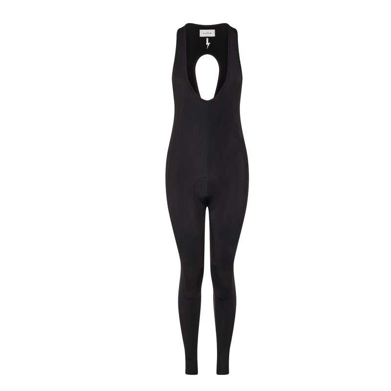 A-full-length-outdoor-lifestyle-photo-featuring-a-young-woman-with-wavy-brown-hair-wearing-a-fitted-long-sleeve-black-jumpsuit- unitard-with-full-length-legs-The- garment-appears-to-have-a-subtle- ribbed-textured-pattern-and-features-a-slightly-scooped-neck- with-layered-detailing-She-is- wearing-oversized-dark-sunglasses- and-black-two-strap-slip-on-sandals-(like-Birkenstocks)-She-is- leaning-against-a-textured-stone- wall-with-a-dark-slatted-fence-above-it-standing-on-a-cobblestone-pavement