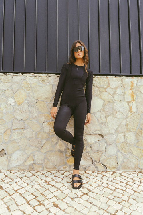 A-full-length-outdoor-lifestyle-photo-featuring-a-young-woman-with-wavy-brown-hair-wearing-a-fitted-long-sleeve-black-jumpsuit- unitard-with-full-length-legs-The- garment-appears-to-have-a-subtle- ribbed-textured-pattern-and-features-a-slightly-scooped-neck- with-layered-detailing-She-is- wearing-oversized-dark-sunglasses- and-black-two-strap-slip-on-sandals-(like-Birkenstocks)-She-is- leaning-against-a-textured-stone- wall-with-a-dark-slatted-fence-above-it-standing-on-a-cobblestone-pavement