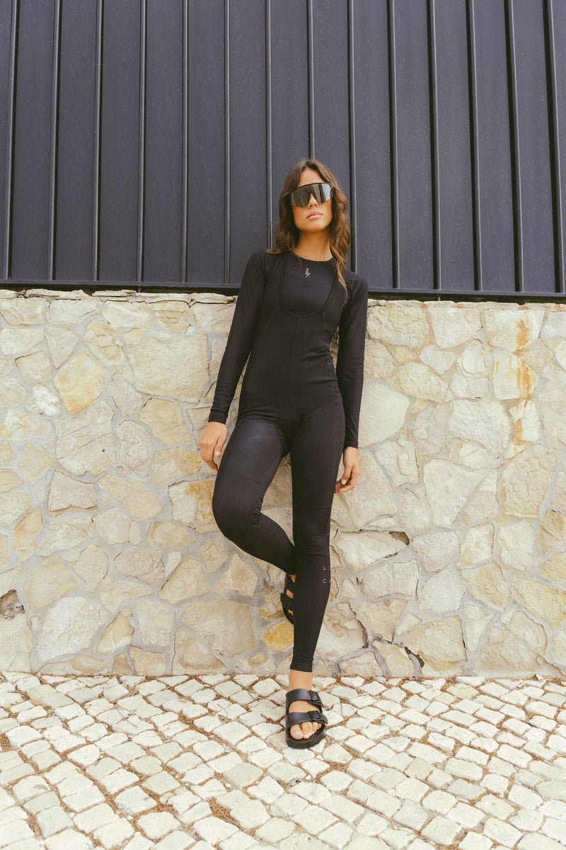 A-full-length-outdoor-lifestyle-photo-featuring-a-young-woman-with-wavy-brown-hair-wearing-a-fitted-long-sleeve-black-jumpsuit- unitard-with-full-length-legs-The- garment-appears-to-have-a-subtle- ribbed-textured-pattern-and-features-a-slightly-scooped-neck- with-layered-detailing-She-is- wearing-oversized-dark-sunglasses- and-black-two-strap-slip-on-sandals-(like-Birkenstocks)-She-is- leaning-against-a-textured-stone- wall-with-a-dark-slatted-fence-above-it-standing-on-a-cobblestone-pavement