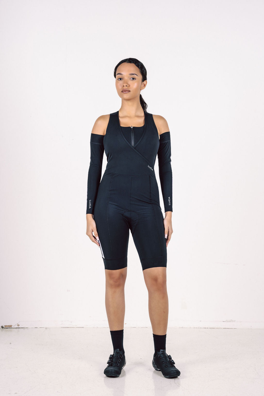 A-full-length-studio-photograph-of-a woman-standing-against-a-white background-wearing-a-navy-blue- cycling-unitard-that-features-a-biker-short-length-The-suit-has-a- sleeveless-crossed-front-V-neck- design-with-an-inne-zippered-layer- and-is-paired-with-detachable- matching-navy-blue-arm-warmers- for-a-cold-shoulder-effect-The-suit- and-arm-warmers-have-white- accent-stripes-on-the-sides-(visible- on-the-legs)-and-the-brand-name- "LUCA"-is-on-the-garment-She-is- wearing-black-cycling-shoe